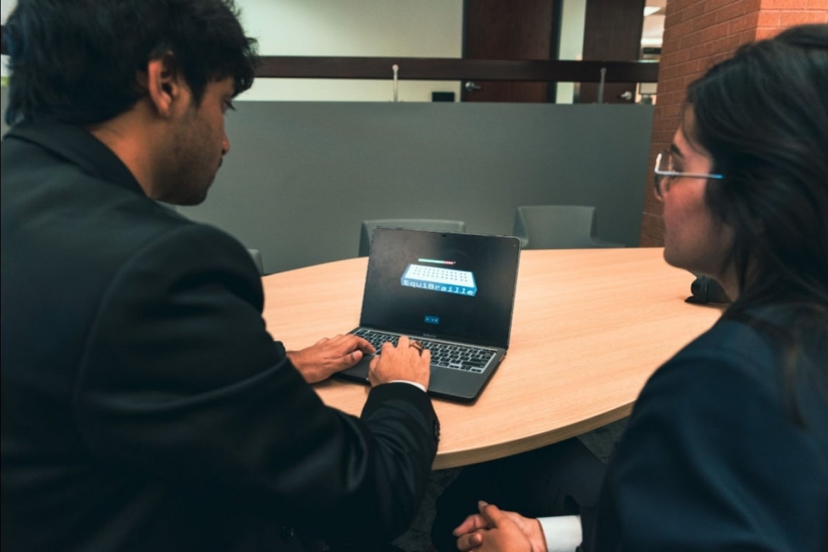 Two students work on a laptop with a 3D render of a board with 