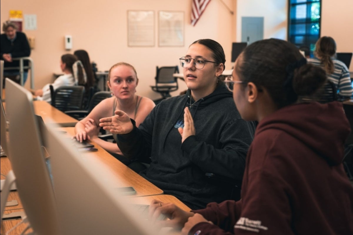 Students sit at a long table with computers, discussing their project.