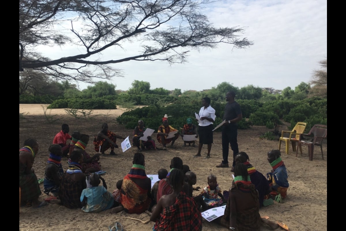 People seated outdoors in a semi-circle around two people standing and speaking.