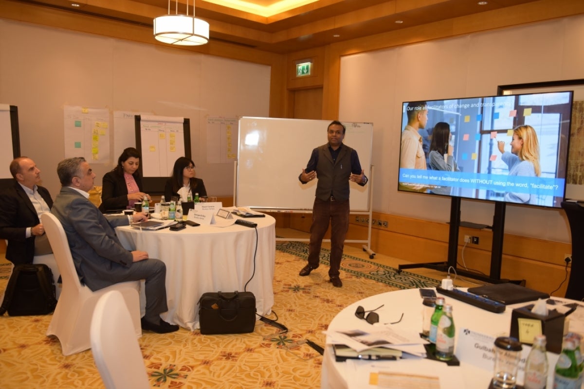 Person standing in front of a room of people sitting at tables in front of a presentation screen.