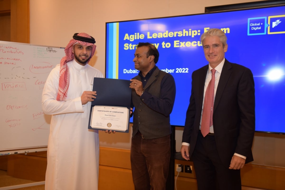 Student receiving their certificate and standing in front of a presentation screen with two other people, one of whom is hanging over the certificate.