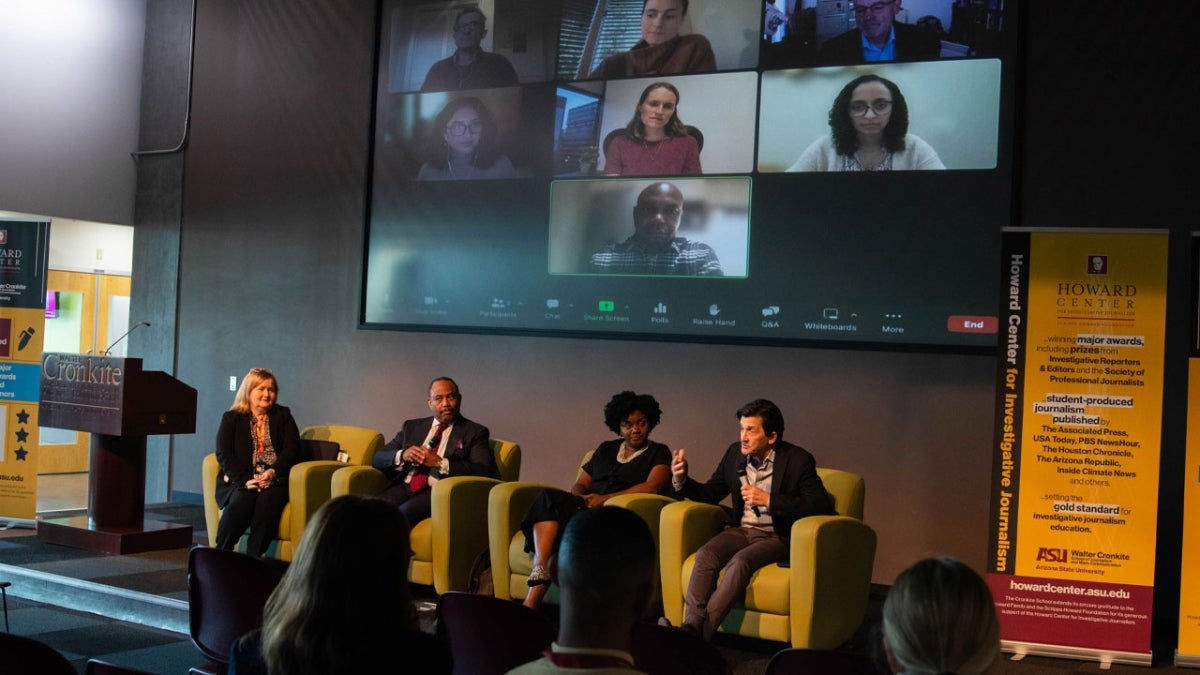 People seated in chairs on a stage with a screen behind them showing various people listening on a Zoom call. One man on stage is speaking into a microphone.