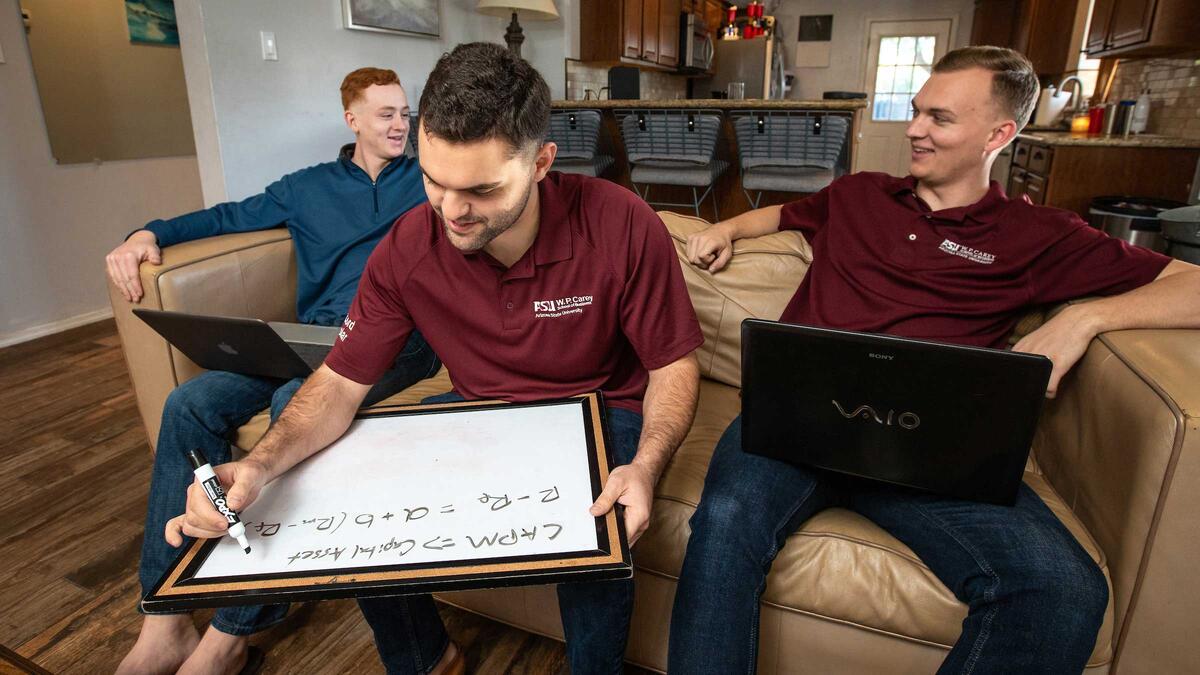 Three roommates sit on a couch working on laptops and a whiteboard