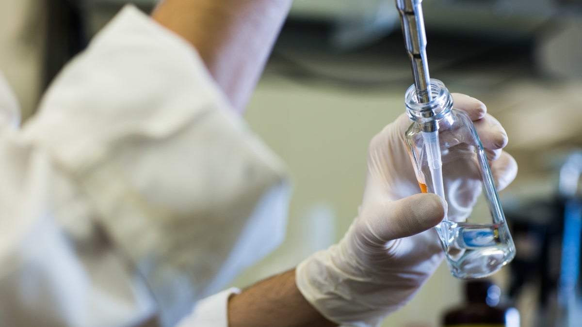 Gloved hands piping liquid into a vial in a lab.