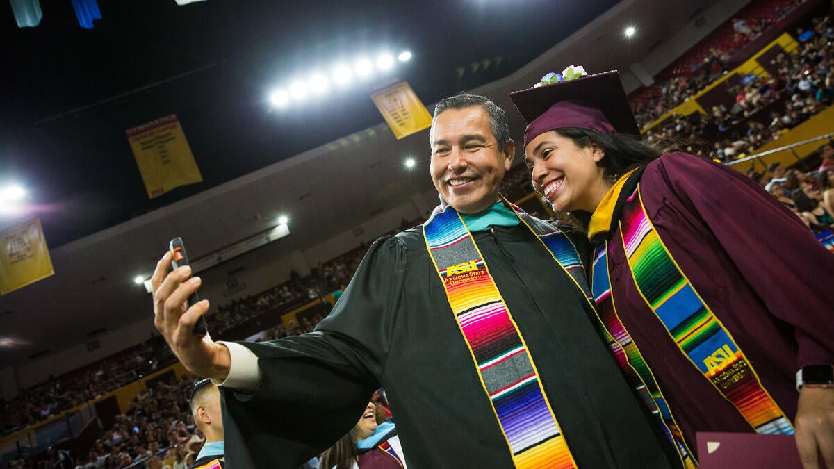 Young student posing with a program director at ASU's Hispanic convocation. Both wear graduation regalia, with the student in a maroon cap and gown and the director in a black gown, and both wear bright, multi-colored stoles.