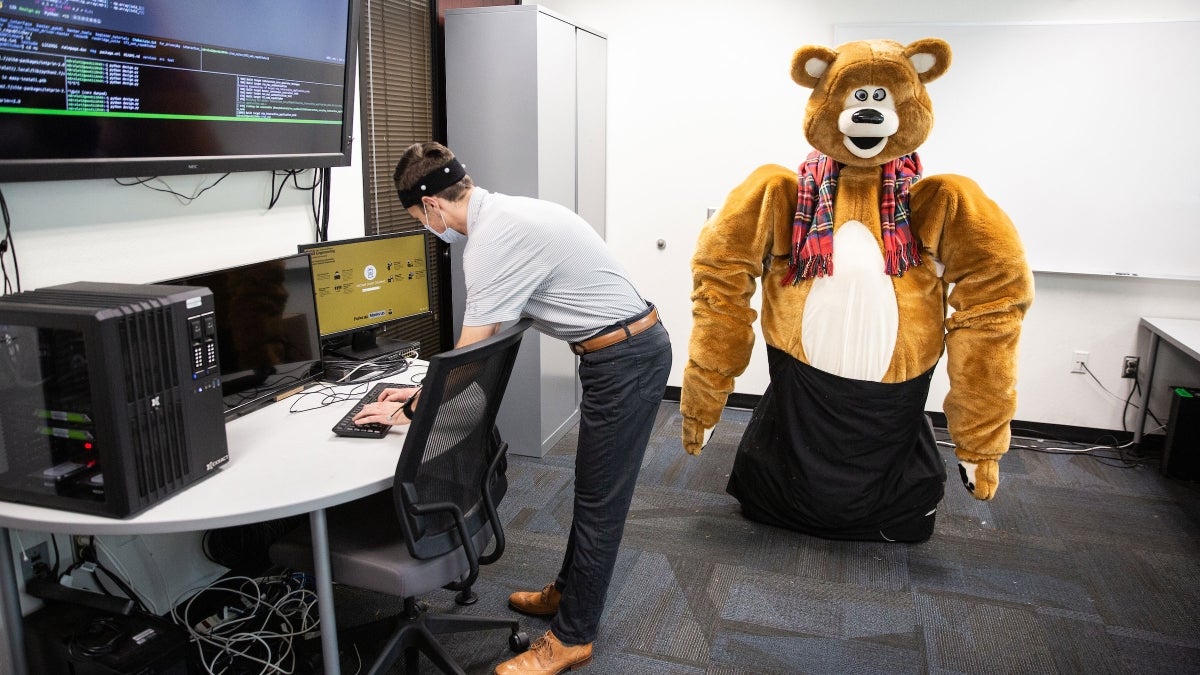 A person works at a computer desk while a large robot made to look like a giant teddy bear stands in the background.