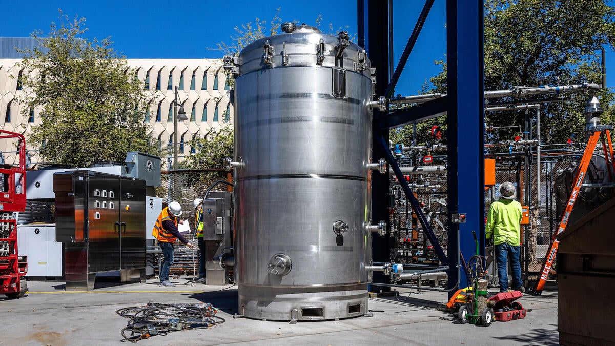 A large metal canister twice as tall as people sits outside surrounded by mechanical equipment 