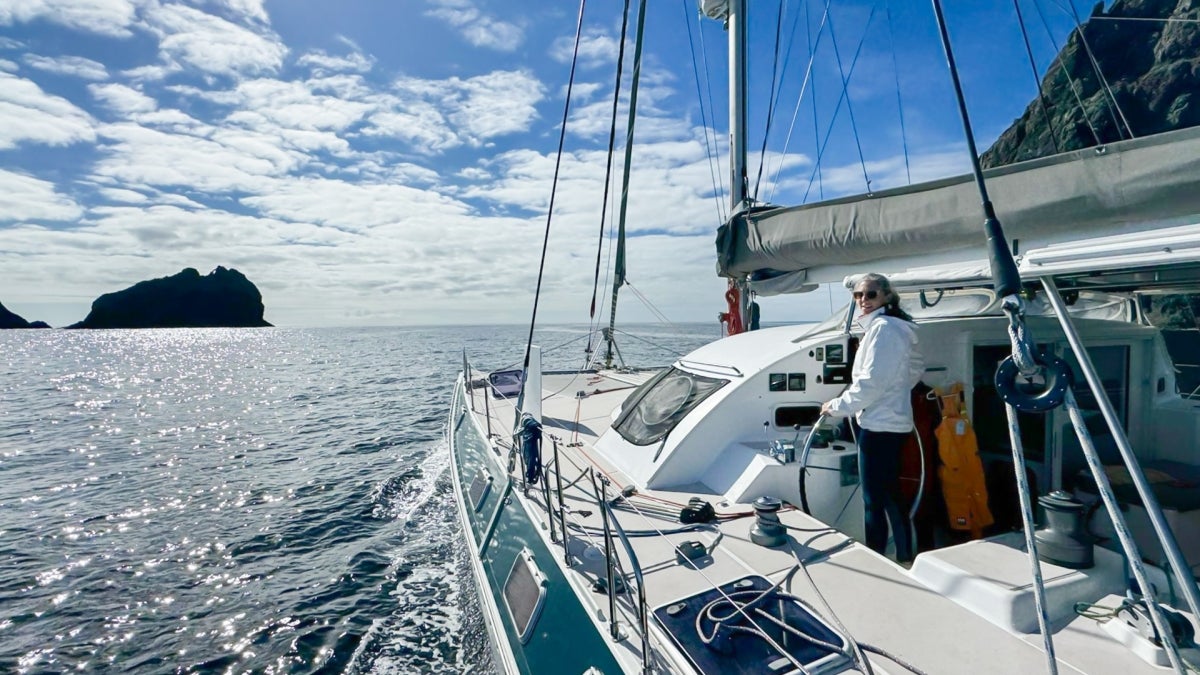 A sailboat on open water with Julia Tizard Hunter steering, rocky landmass in the distance, and clouds in the sky.