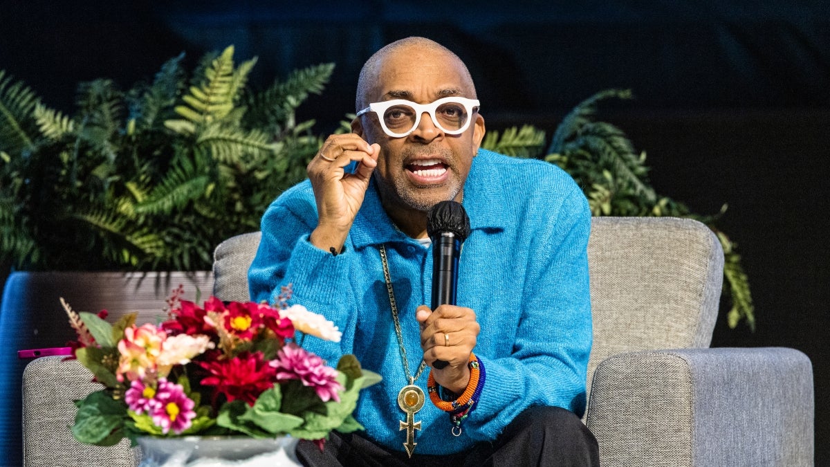 Filmmaker Spike Lee speaks into a microphone while siting on a couch on stage during a lecture event