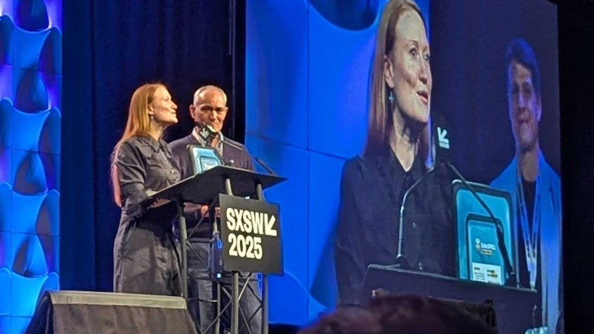 A man and a woman stand on a large stage speaking into a microphone at a podium.