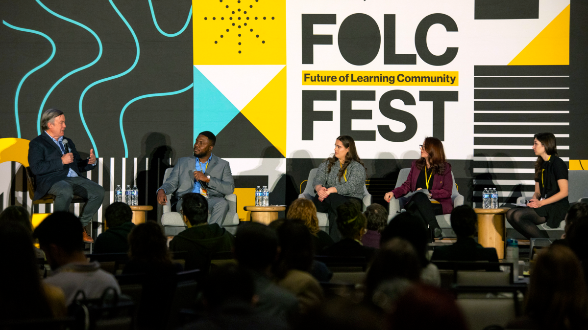 People seated in chairs on a stage listen as one of them speaks into a microphone and an audience watches.