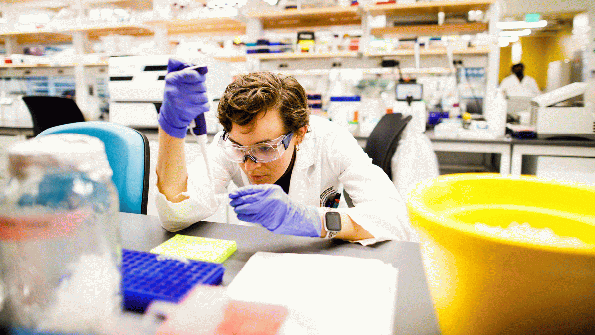 Student in lab gear hunches over lab table to fill vials with a pipette