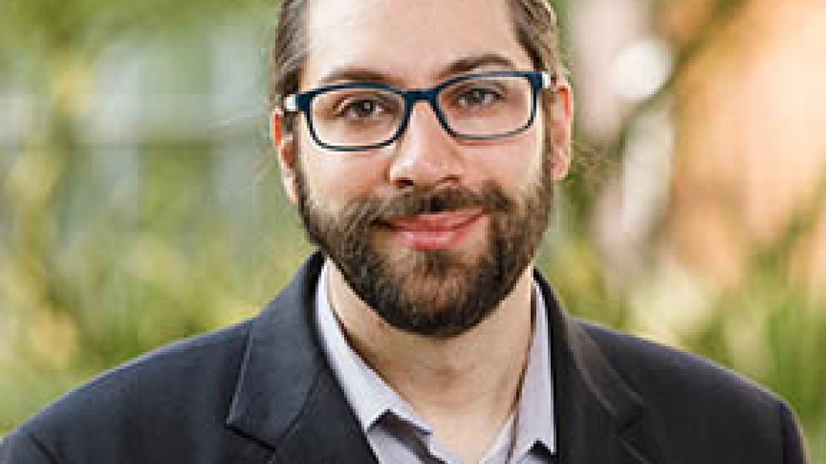 ASU Assistant Professor Sam Teitelbaum is pictured from the chest up, smiling, in front of a blurry outdoor background.