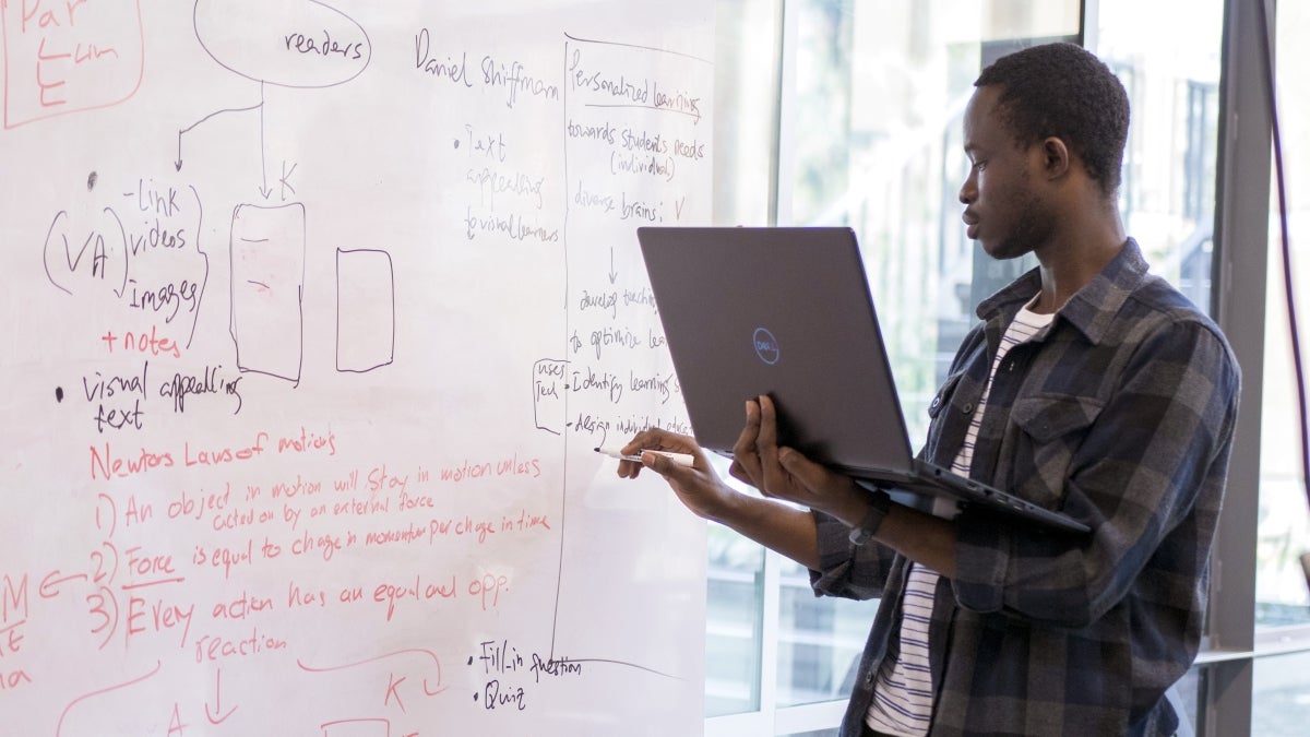 A student holds a laptop and writes on a dry erase board.