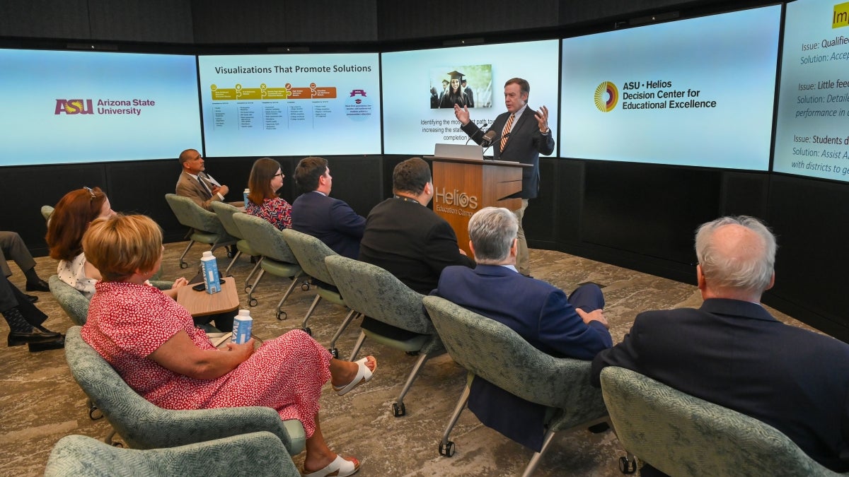 A man speaks at a lectern to a small audience in room with large display screens on every wall