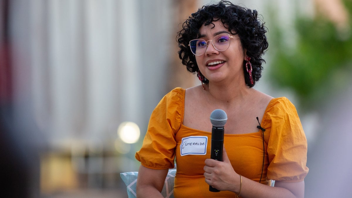 Woman with glasses and short, curly brown hair speaking into a microphone.