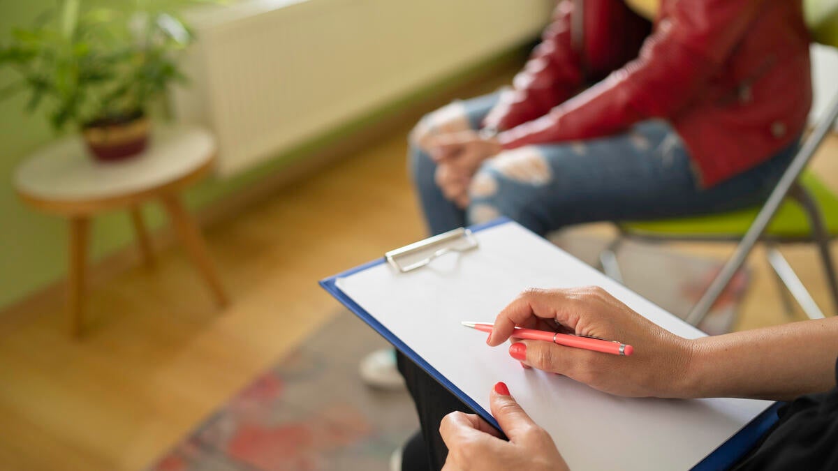 Hands holding a clipboard next to a seated young person whose face is not seen.