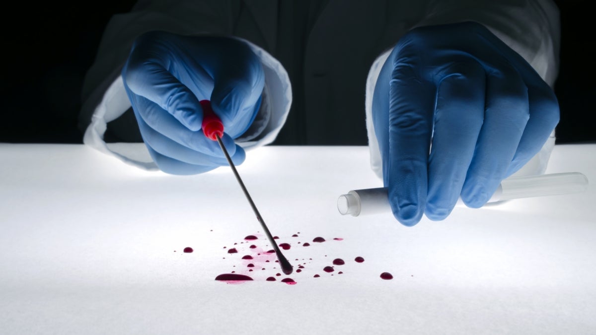 Blue-gloved hands hold a swab and a test tube over red droplets.