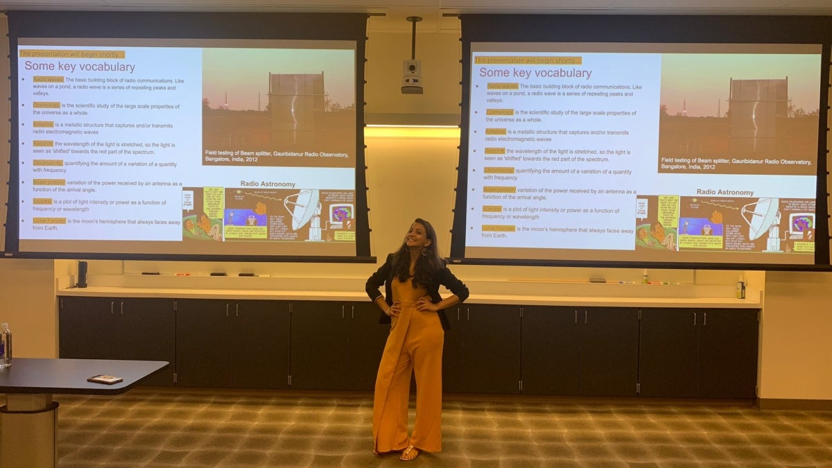 Nivedita Mahesh stands in the front of a classroom, with a presentation displayed on two screens behind her.