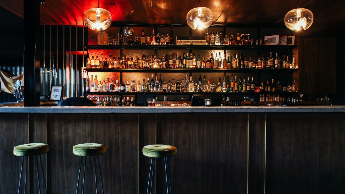 An empty bar with three green barstools in front of it and shelves full of liquor behind it.