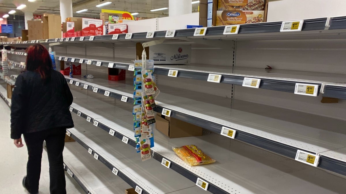 Person seen from the back, walking down an aisle in a grocery store with empty shelves.