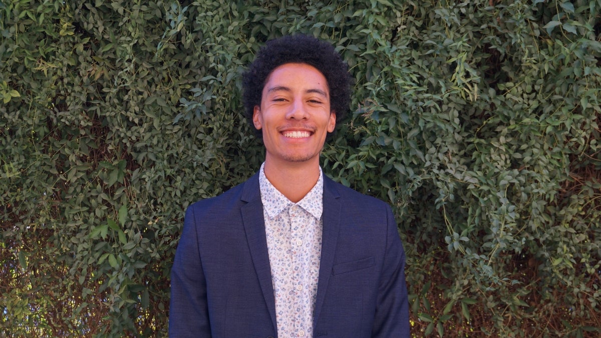 Man wearing collared shirt and blazer smiles in front of wall of greenery