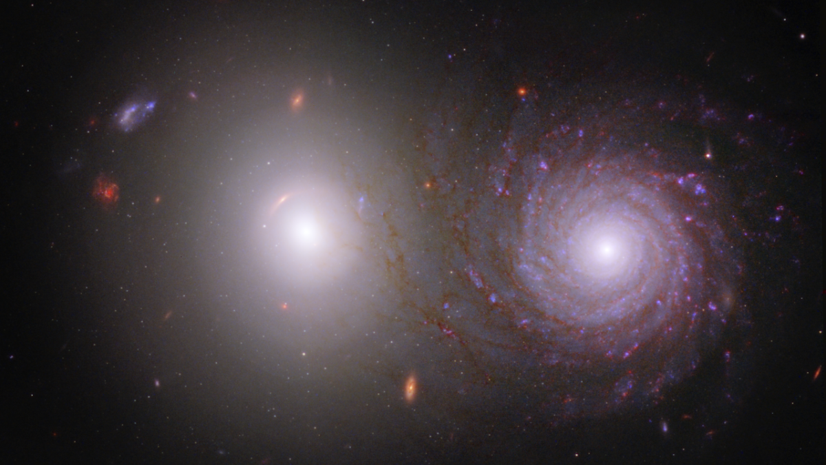 The majority of the image shows the black background of space. Two large, very bright galaxies dominate the center of the image. The elliptical galaxy at left is extremely bright at its circular core, with dimmer white light extending to its transparent c