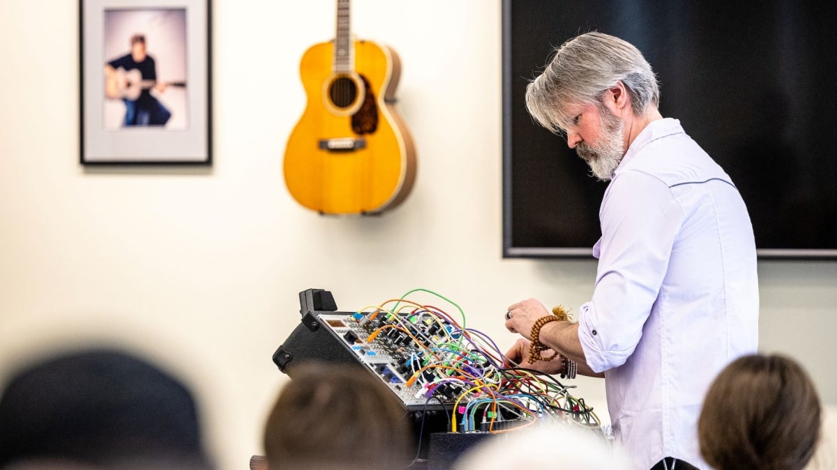 Man standing at a board with several wires poking out of it in front of a wall with a guitar hanging on it.