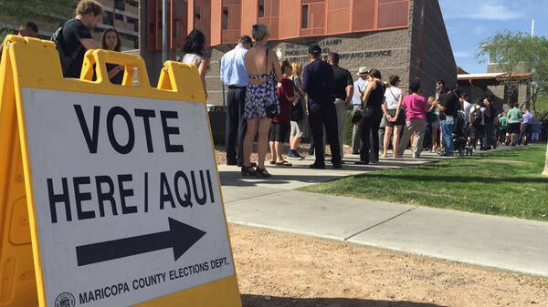 Sign reading "Vote here/aqui" in the foreground and a line of people in the background.