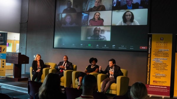 People seated in chairs on a stage with a screen behind them showing various people listening on a Zoom call. One man on stage is speaking into a microphone.