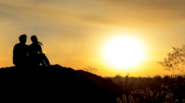 A silhouetted man and women sit on a mountain top looking out at a golden sunset.