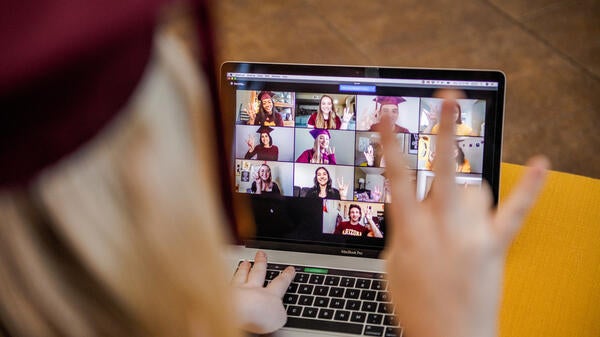 A student wearing a graduation cap makes the forks up gesture at a Zoom screen featuring other graduates