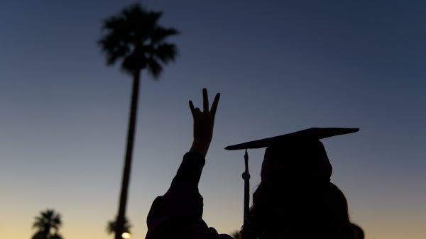 silhouette of student in cap and gown making a pitchfork sign with palm tree behind them