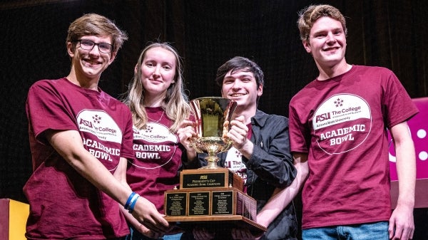 Four college students hold a large trophy after winning the Academic Bowl