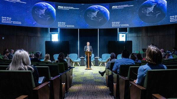 Man speaking behind a lectern at an event with TV screens displaying Earth behind him.