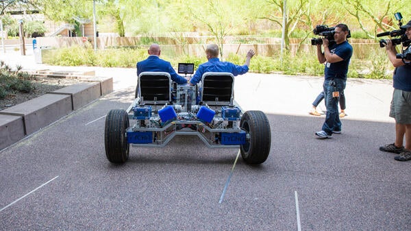 Two men in a lunar rover drive away on a sidewalk.