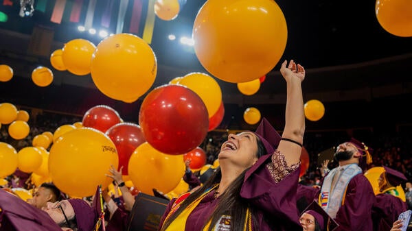 A woman in a graduation cap and gown smiles toward the ceiling as giant balloons drop down onto the graduates