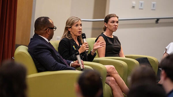 Three people seated on a stage as one of them speaks into a microphone to an unseen audience.