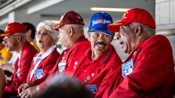 Older men in Tuskegee Airmen hats chat while sitting in stadium seats.