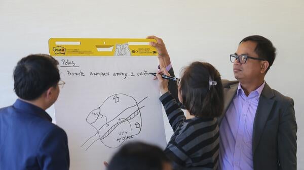 A woman writes on an easel, "Policy = Promote awareness among 2 countries". A map of a city bisected by a river with mining and agriculture sectors has also been drawn.