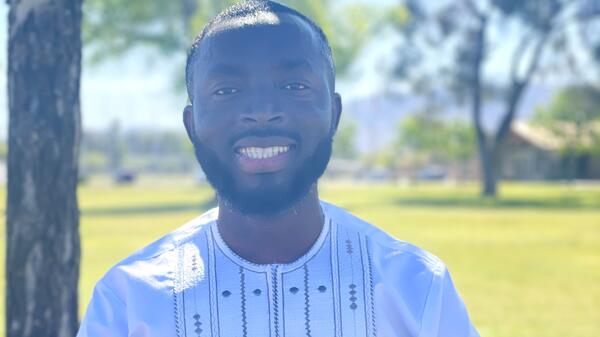 A smiling man dressed in a white shirt stands near a tree on a sunny day.