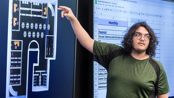 A young man points to a large screen in front of a classroom