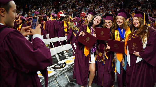 Four people in graduation regalia stand together for a photo while another takes the photo on their phone.