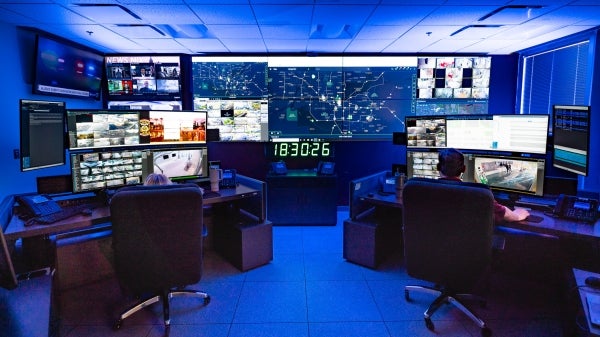 People sit at desks full of several monitors displaying footage of activity taking place around a college campus.