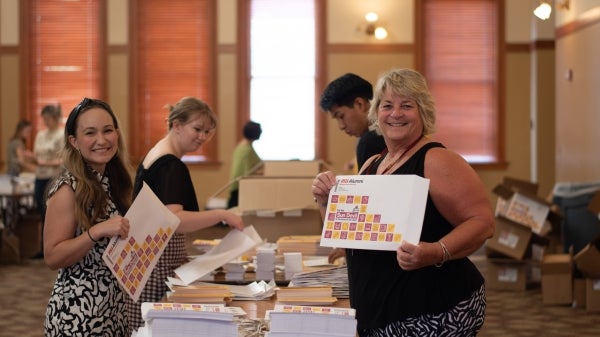 People working at a table filled with printed materials in a well-lit room, two women in the foreground are smiling at the camera.