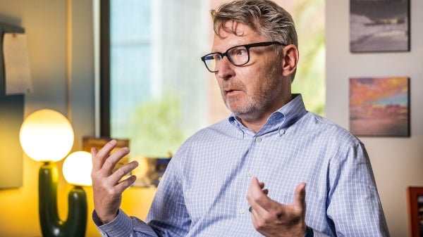 Man with salt and pepper hair and black glasses wearing a blue and white checked shirt talks in office