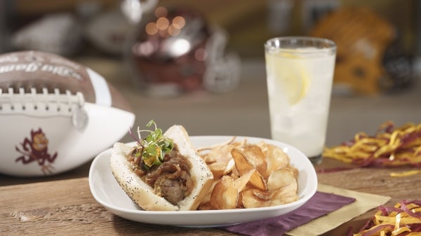 A plate with bratwurst and chips sits on a table next to an ASU football and a glass of water