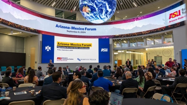 Woman speaking behind a lectern in front of a large screen that reads "Arizona-Mexico Forum" as a large audience looks on.