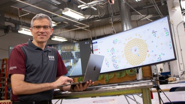 Man holding a laptop while leaning on a table with a desktop computer on it displaying various data.