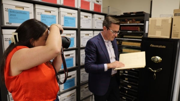 Woman photographing a man standing next to a filing cabinet while looking at a manila folder.